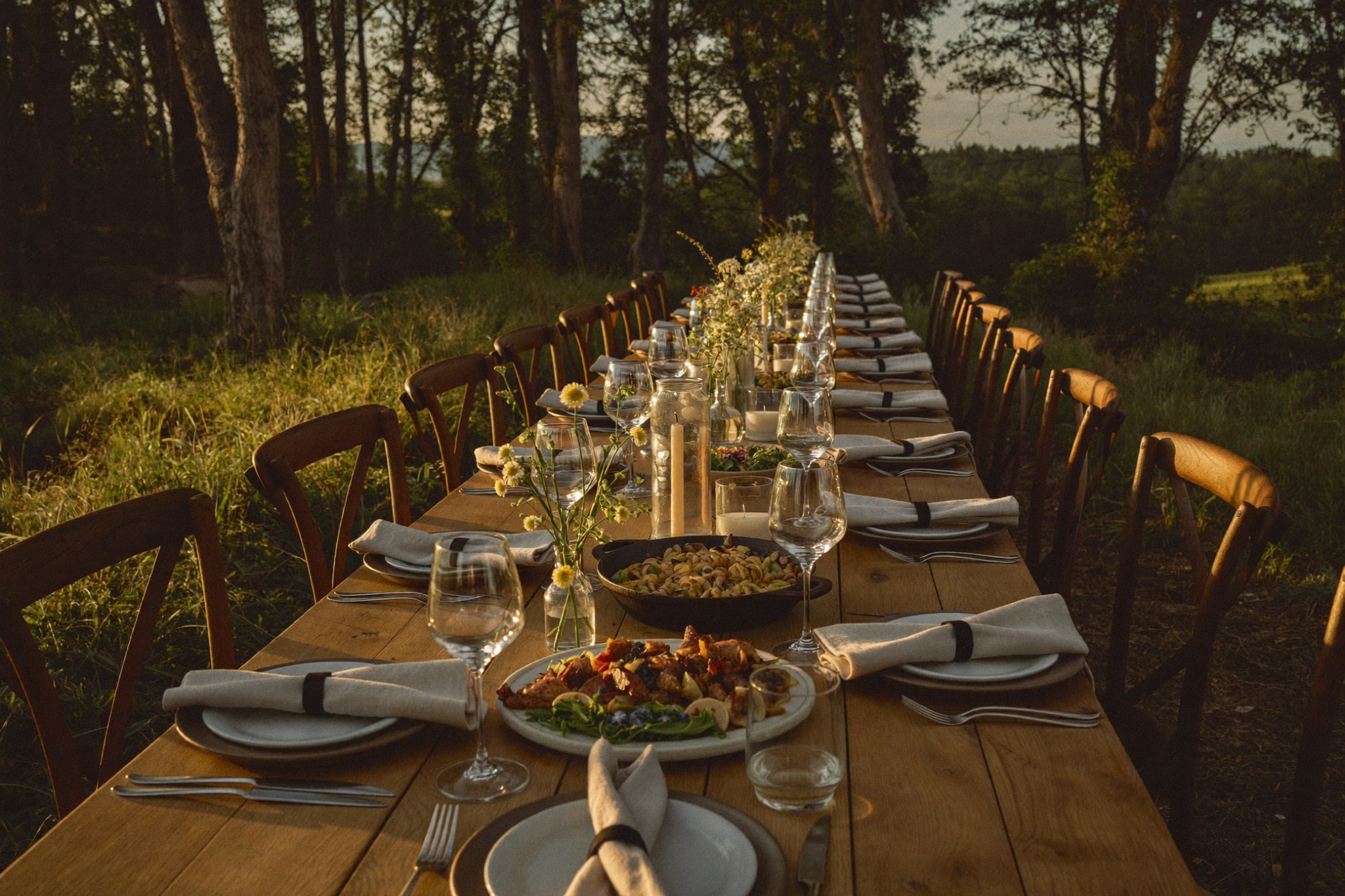Long oak table set for a fire-cooked dinner at the forest edge